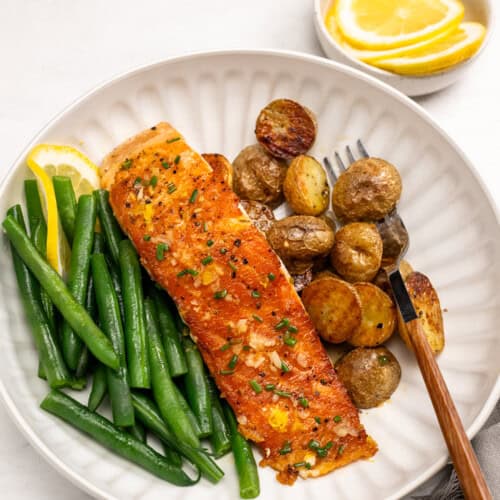 A plate with a cooked salmon fillet garnished with chives, green beans, and roasted baby potatoes, served with lemon wedges. A small bowl of lemon slices is in the background.