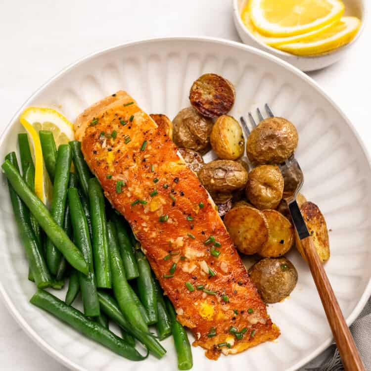 A plate with a cooked salmon fillet garnished with chives, green beans, and roasted baby potatoes, served with lemon wedges. A small bowl of lemon slices is in the background.