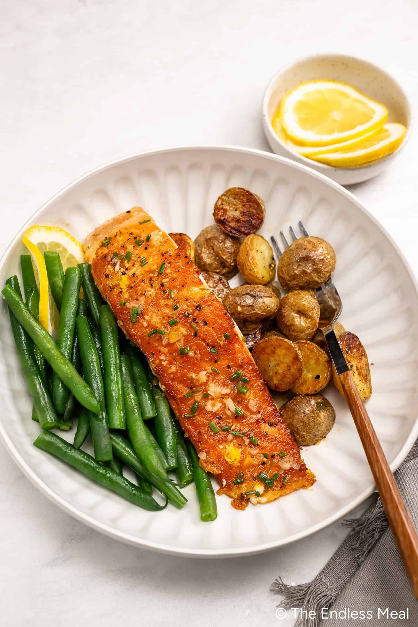 A plate with a cooked salmon fillet garnished with chives, green beans, and roasted baby potatoes, served with lemon wedges. A small bowl of lemon slices is in the background.