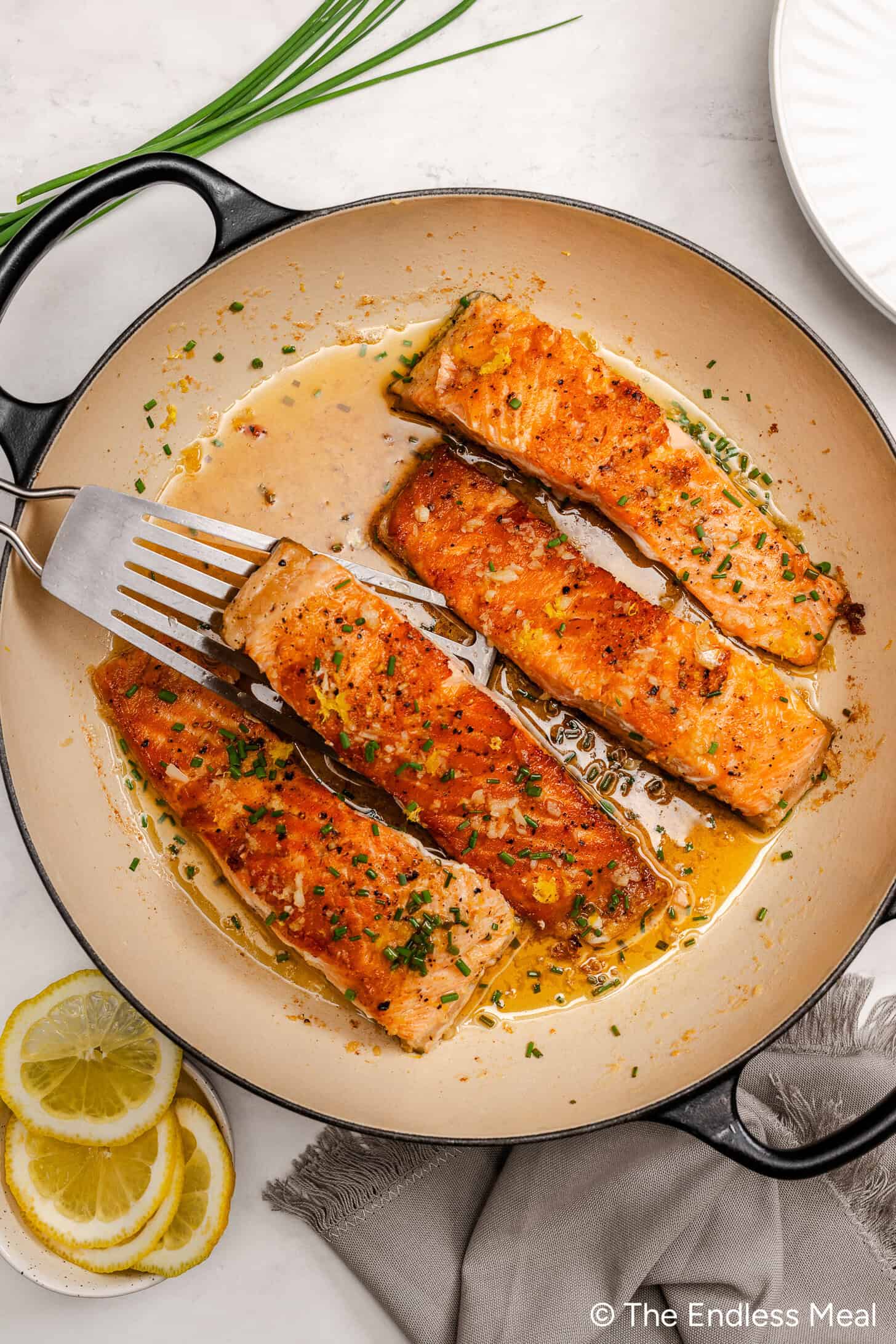 Four cooked salmon fillets in a pan with a light lemon butter sauce, garnished with chopped chives. A spatula lifts one fillet, and lemon slices and extra chives are nearby—a simple and delicious Lemon Butter Salmon recipe.