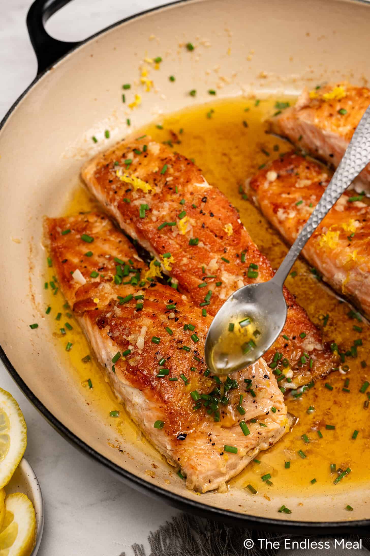 Salmon fillets in a skillet with lemon butter sauce being spooned over the top, garnished with chopped chives and lemon zest for a bright, flavorful Lemon Butter Salmon.