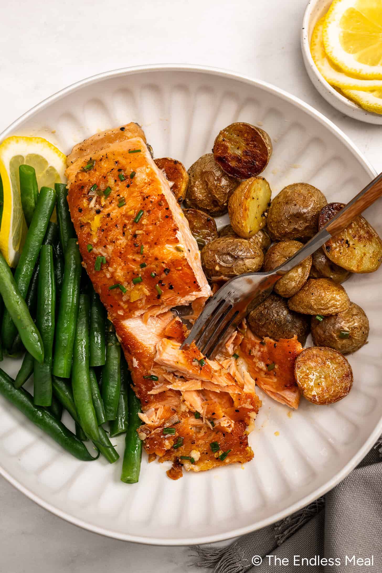 A plate with a cooked lemon butter salmon fillet garnished with chives, roasted baby potatoes, green beans, and a lemon slice. A fork is flaking the salmon.