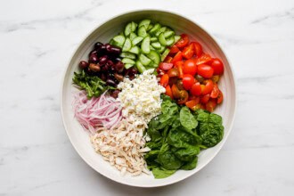 Image for recipe instruction: A bowl containing Mediterranean Orzo Salad, with spinach, shredded chicken, sliced red onions, black olives, cucumber, cherry tomatoes, feta cheese, and fresh herbs arranged in separate sections.