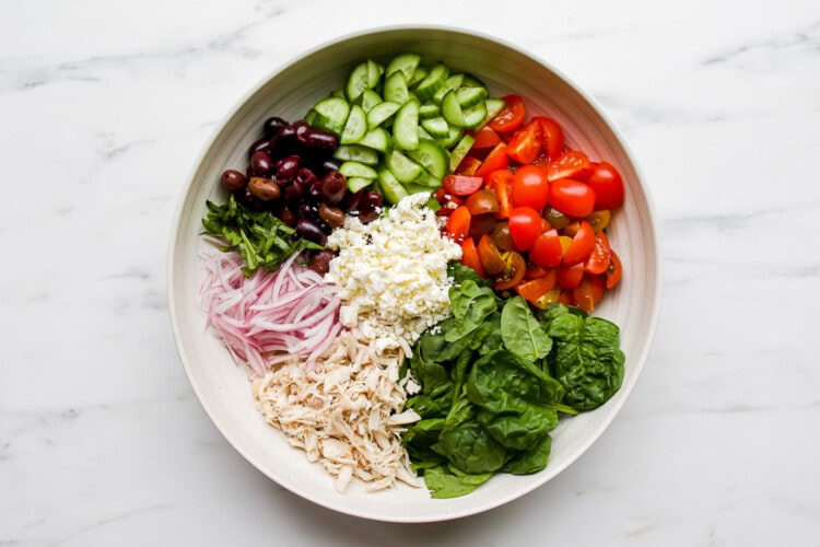 Image for recipe instruction: A bowl containing Mediterranean Orzo Salad, with spinach, shredded chicken, sliced red onions, black olives, cucumber, cherry tomatoes, feta cheese, and fresh herbs arranged in separate sections.