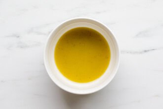 Image for recipe instruction: A white bowl filled with yellow soup sits on a white marble surface, next to a refreshing Mediterranean Orzo Salad.