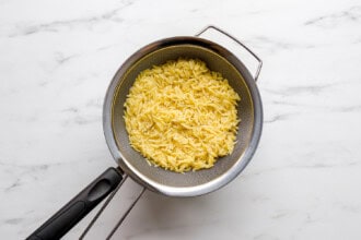 Image for recipe instruction: A metal strainer containing cooked orzo pasta, perfect for preparing a refreshing Mediterranean Orzo Salad, sits on a white marble surface.