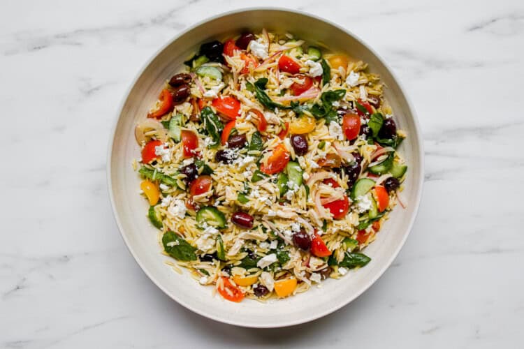 Image for recipe instruction: A bowl of Mediterranean Orzo Salad with cherry tomatoes, cucumbers, spinach, olives, and crumbled cheese on a white marble surface.