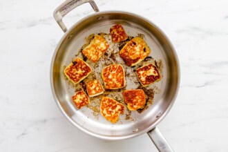 A stainless steel pan on a marble surface with several pieces of fried halloumi cheese being browned, perfect as a tasty topping for your favorite tabbouleh recipe.