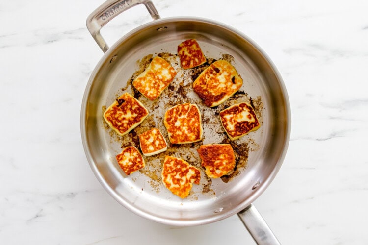 A stainless steel pan on a marble surface with several pieces of fried halloumi cheese being browned, perfect as a tasty topping for your favorite tabbouleh recipe.
