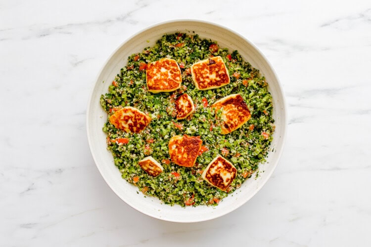A white bowl filled with broccoli tabbouleh salad topped with several pieces of golden-brown fried halloumi, placed on a white marble surface.