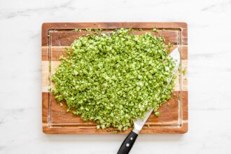 Finely chopped broccoli on a wooden cutting board with a chef’s knife, ready to be used in a fresh Broccoli Tabbouleh recipe, all set atop a white marble surface.
