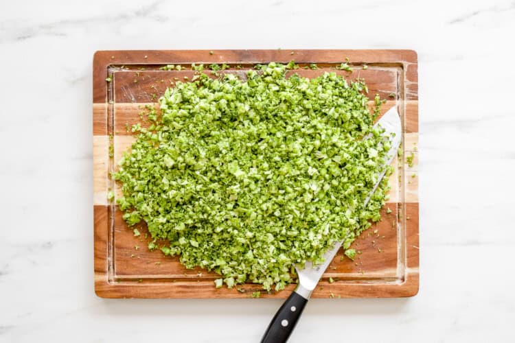 Finely chopped broccoli on a wooden cutting board with a chef’s knife, ready to be used in a fresh Broccoli Tabbouleh recipe, all set atop a white marble surface.