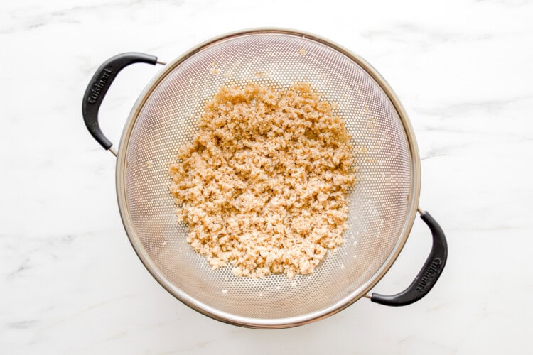 Cooked bulgar draining in a metal mesh strainer with black handles on a white marble surface, perfect for adding to your favorite Broccoli Tabbouleh or next Tabbouleh Recipe.