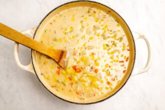 image for recipe instruction: A pot of simple corn chowder with carrots, potatoes, and celery, stirred with a wooden spatula on a light marble surface; this creamy corn chowder uses corn stock for extra flavor.