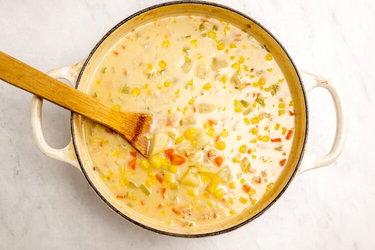 image for recipe instruction: A pot of simple corn chowder with carrots, potatoes, and celery, stirred with a wooden spatula on a light marble surface; this creamy corn chowder uses corn stock for extra flavor.
