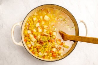 image for recipe instruction: A pot of creamy corn chowder with potatoes, carrots, celery, and corn is being stirred with a wooden spoon on a white surface.