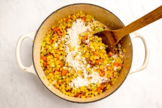 image for recipe instruction: A pot containing chopped carrots, celery, and onions being stirred with a wooden spoon, with a layer of flour sprinkled on top—perfect for starting a simple corn chowder or adding flavor to your favorite corn stock-based soups.