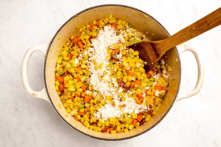 image for recipe instruction: A pot containing chopped carrots, celery, and onions being stirred with a wooden spoon, with a layer of flour sprinkled on top—perfect for starting a simple corn chowder or adding flavor to your favorite corn stock-based soups.