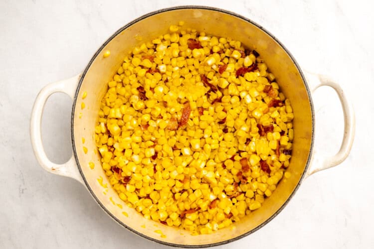 image for recipe instruction: A large white pot filled with cooked corn kernels and bacon on a light marble surface, perfect for starting a simple corn chowder.