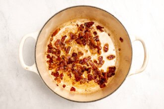 image for recipe instruction: Chopped bacon pieces cooking and browning in a large white Dutch oven on a light countertop, creating the perfect savory base for Creamy Corn Chowder or simple corn stock.