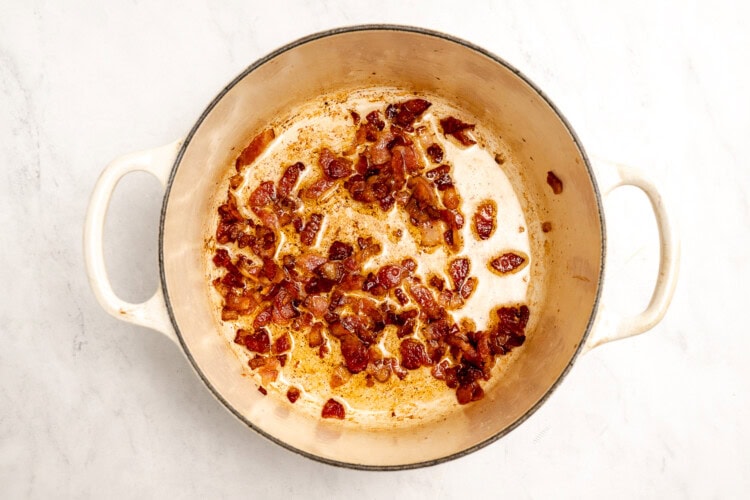 image for recipe instruction: Chopped bacon pieces cooking and browning in a large white Dutch oven on a light countertop, creating the perfect savory base for Creamy Corn Chowder or simple corn stock.