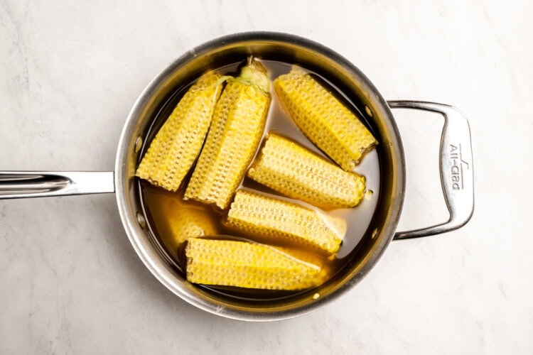 image for recipe instruction: A stainless steel saucepan filled with corn cobs simmering in yellow liquid, perfect for making a simple corn chowder or flavorful corn stock, sits on a white marble surface.
