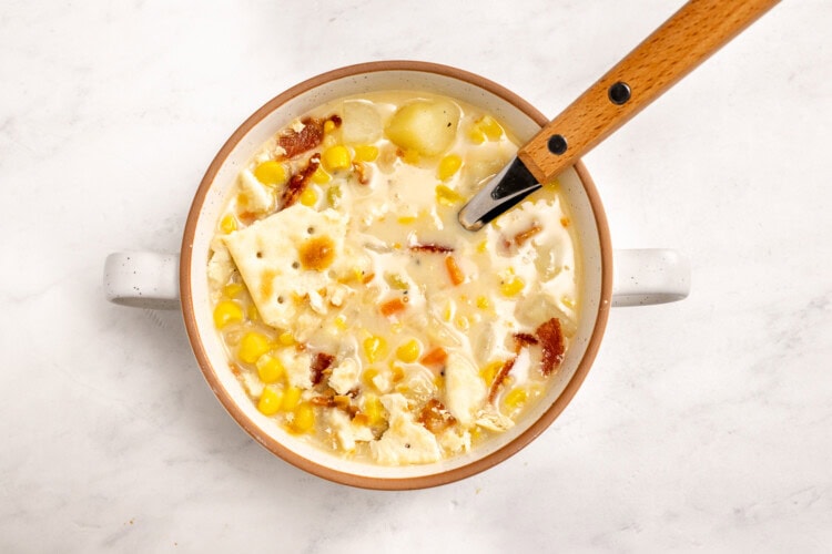 image for recipe instruction: A bowl of Simple Corn Chowder with creamy corn stock, potato chunks, bacon pieces, and saltine crackers, with a wooden-handled spoon resting inside.