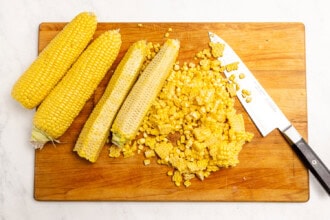 image for recipe instruction: Two whole corn cobs, two stripped cobs, and corn kernels rest on a wooden cutting board next to a knife—perfect ingredients for making creamy corn chowder or preparing a fresh corn stock.