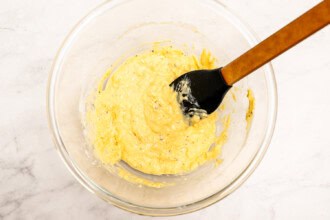 Image for recipe instruction: A glass bowl containing a yellow, creamy mixture for a delicious Corn Fritters recipe is being stirred with a black spatula on a light countertop.