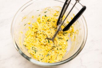 Image for recipe instruction: A glass bowl containing a yellow corn mixture for corn fritters with chopped herbs and a metal scoop resting inside, on a white surface.