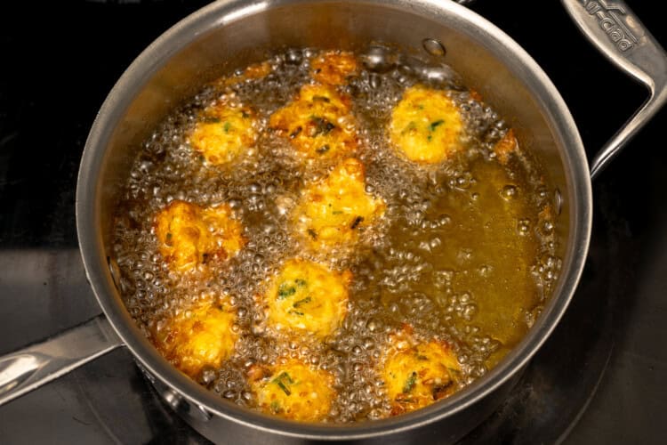 Image for recipe instruction: Golden corn fritters are being deep-fried to perfection in hot oil in a stainless steel pan on a stovetop.