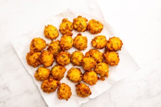 Image for recipe instruction: A group of golden-brown corn fritters arranged on a white paper towel, placed on a white marble surface.