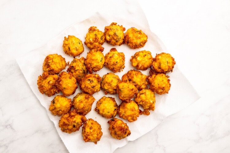 Image for recipe instruction: A group of golden-brown corn fritters arranged on a white paper towel, placed on a white marble surface.