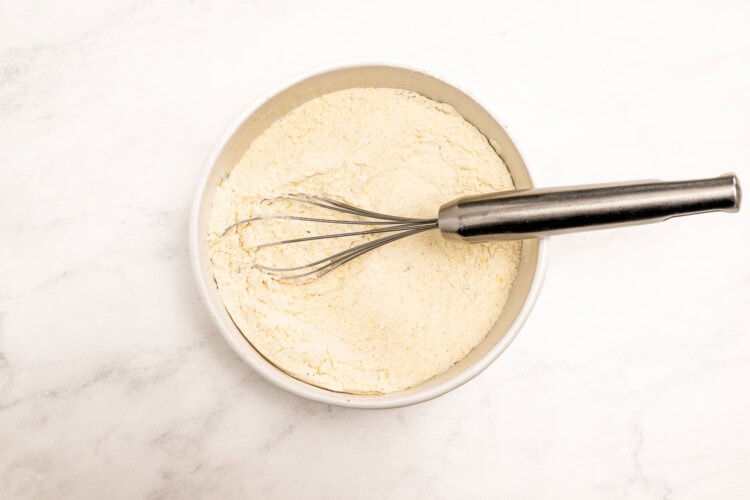 Image for recipe instruction: A metal whisk rests in a white bowl filled with flour on a white marble surface, ready to mix batter for delicious corn fritters.