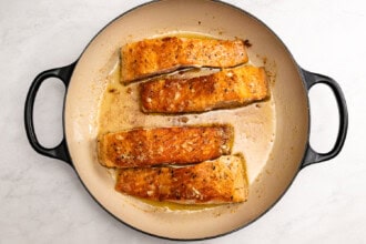 Image for recipe instruction: Four cooked Lemon Butter Salmon fillets in a round, cream-colored skillet with a light sauce and chives, viewed from above on a white surface.