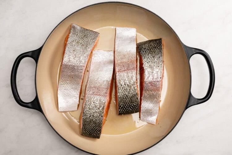Image for recipe instruction: Four raw salmon fillets with skin on are arranged in a large cream-colored pan with black handles, ready to become a delicious Lemon Butter Salmon, set on a white surface.