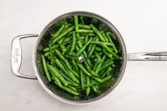 Image for recipe instruction: Green beans in a metal steamer basket inside a saucepan, viewed from above on a white surface—perfect as a fresh side for your favorite Lemon Butter Salmon.