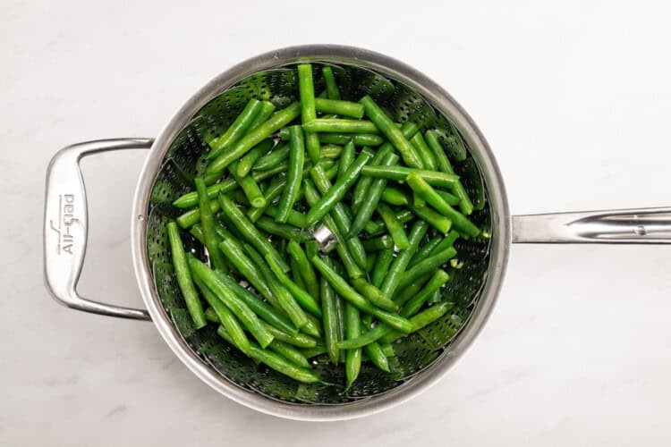 Image for recipe instruction: Green beans in a metal steamer basket inside a saucepan, viewed from above on a white surface—perfect as a fresh side for your favorite Lemon Butter Salmon.