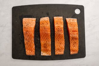 Four raw salmon fillets, seasoned with salt and pepper and ready for a lemon butter salmon recipe, are arranged in a row on a black cutting board.