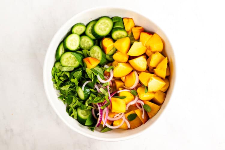 Image for recipe instruction: A white bowl filled with sliced cucumbers, chopped yellow peaches, red onion, and fresh green herbs on a white surface—this vibrant Cucumber Peach Salad is perfect drizzled with a hot honey vinaigrette.