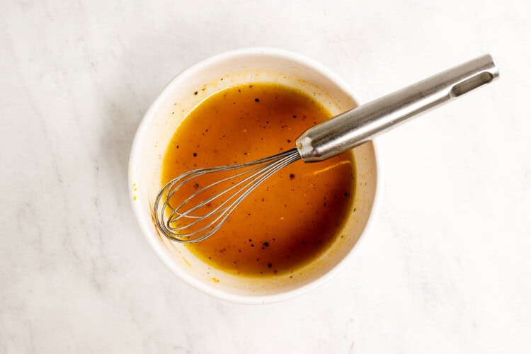 Image for recipe instruction: A metal whisk rests in a white bowl filled with a brown, seasoned Hot Honey Vinaigrette, set on a light marble surface—perfect for drizzling over a fresh Peach Salad.