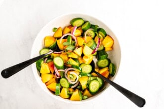 Image for recipe instruction: A bowl of fresh Peach Salad with sliced cucumbers, yellow tomatoes, red onion, and mint, finished with a hot honey vinaigrette and two black utensils placed inside.