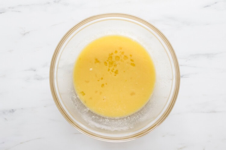 A glass bowl containing a melted butter mixture sits on a white marble surface.
