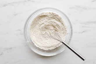 A glass bowl filled with flour and a metal whisk on a white marble surface, ready for preparing strawberry rhubarb muffin batter.