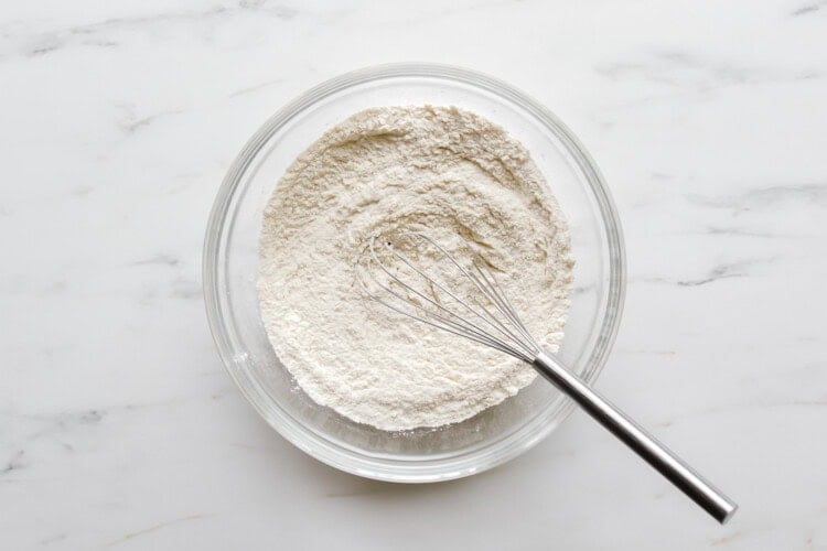 A glass bowl filled with flour and a metal whisk on a white marble surface, ready for preparing strawberry rhubarb muffin batter.