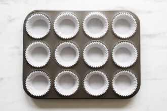 A 12-cup muffin tin with each cup lined with a white paper baking liner, perfect for baking this strawberry rhubarb muffin recipe, placed on a white marble surface.