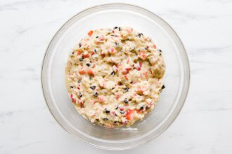 A glass bowl filled with strawberry rhubarb muffin batter, dotted with chopped fruits and dark specks, sits on a white marble surface.