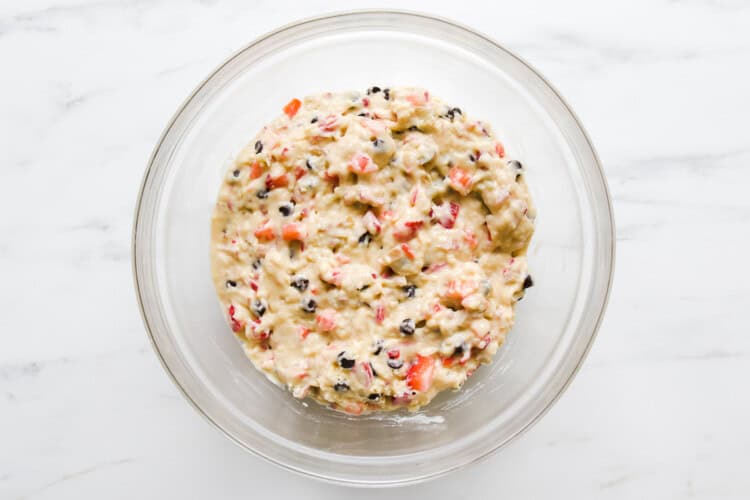 A glass bowl filled with strawberry rhubarb muffin batter, dotted with chopped fruits and dark specks, sits on a white marble surface.