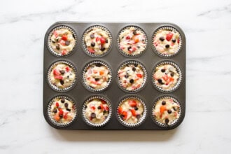 A muffin tin with 12 paper liners filled with unbaked strawberry rhubarb muffin batter, dotted with visible strawberries and chocolate chips, rests on a white countertop.