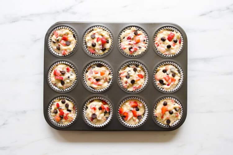 A muffin tin with 12 paper liners filled with unbaked strawberry rhubarb muffin batter, dotted with visible strawberries and chocolate chips, rests on a white countertop.